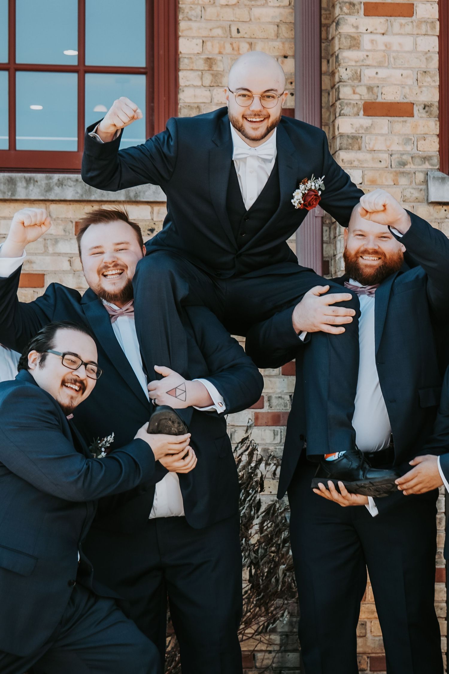 Groomsmen in black suits strike playful poses against a brick wall for fun wedding party photos.