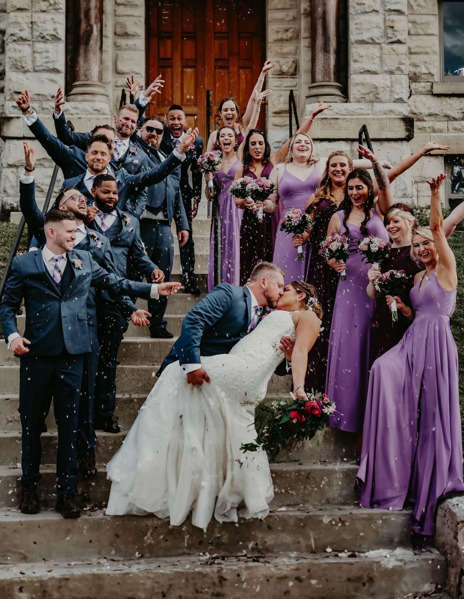 Wedding party in purple bridesmaid dresses and dark suits celebrate joyfully on stone church steps.
