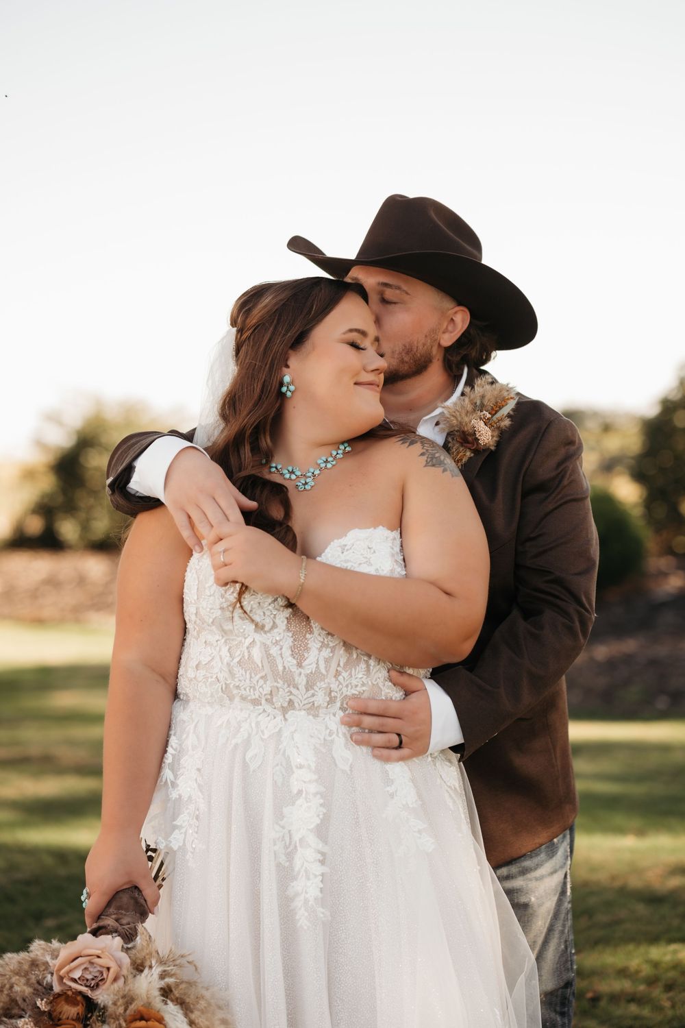 A romantic wedding moment with a bride in white lace dress and groom in brown western attire embracing outdoors at sunset.