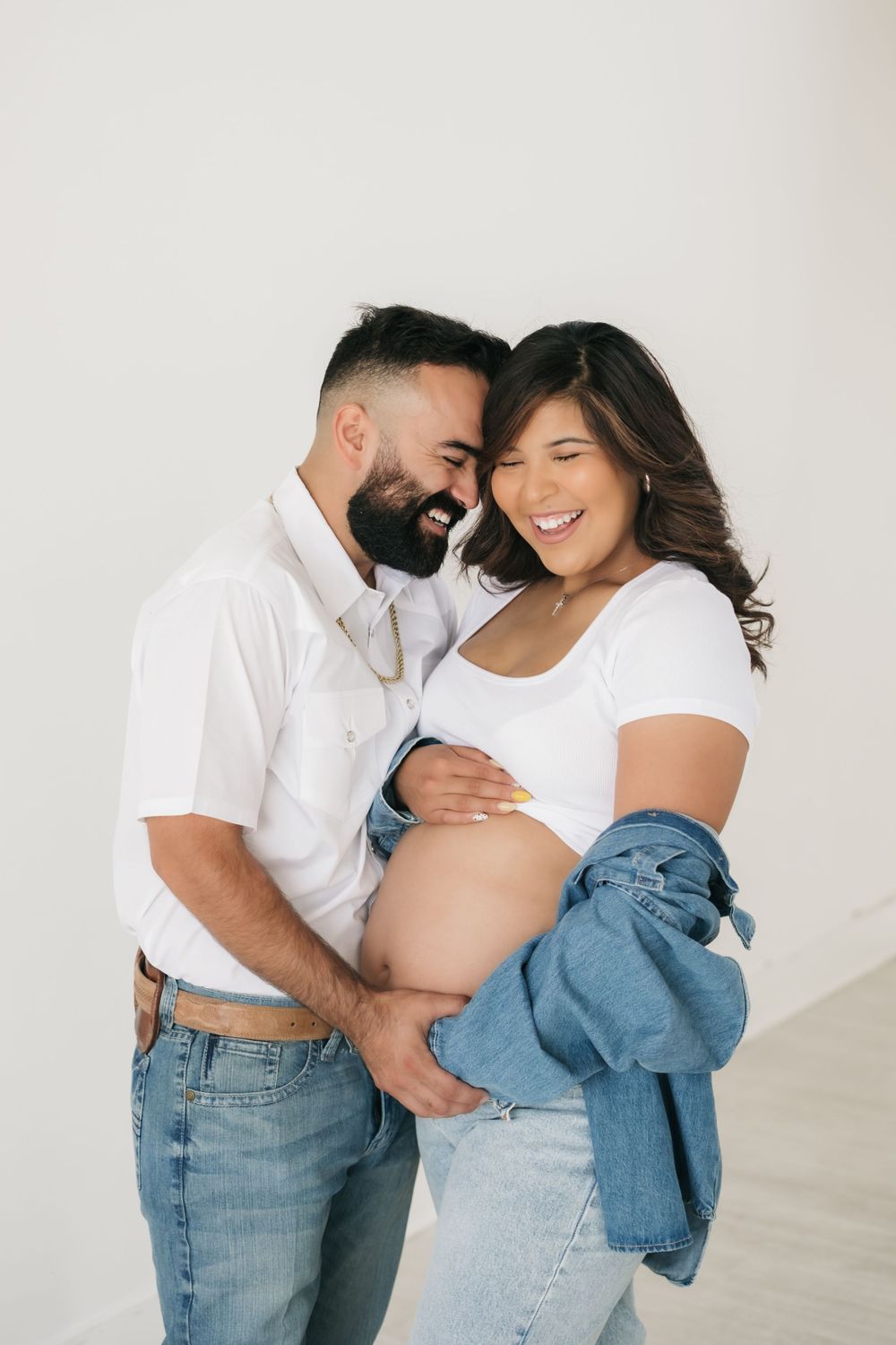 A couple wearing matching white tops and blue jeans embrace intimately in a sweet maternity photoshoot against a white backdrop.