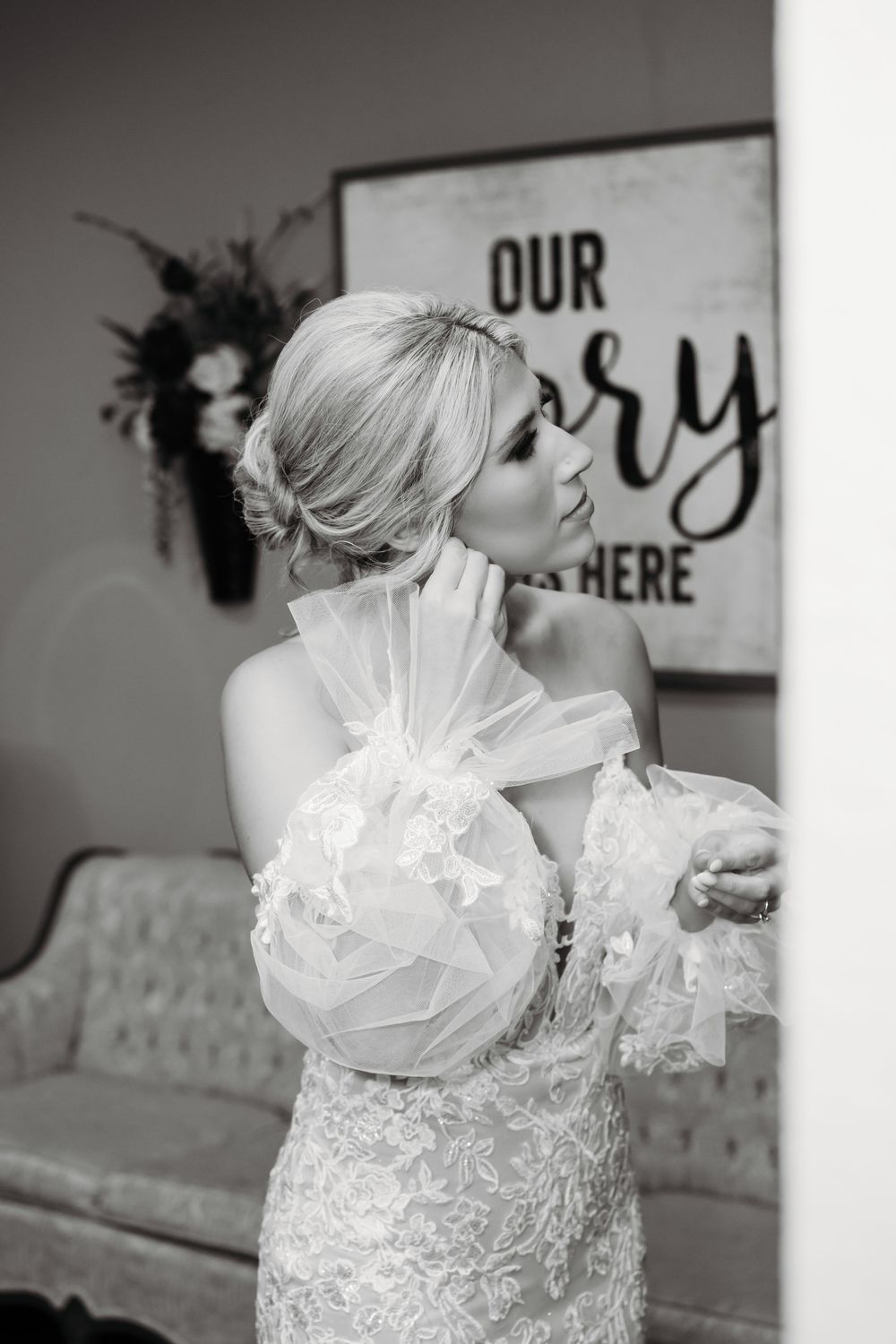 A black and white photo of a bride in a lace wedding dress with sheer ruffled sleeves in an elegant interior setting.
