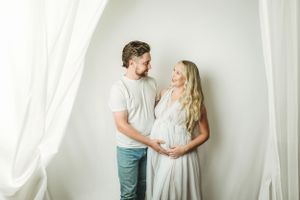 Expecting couple poses lovingly together in a bright white studio setting.