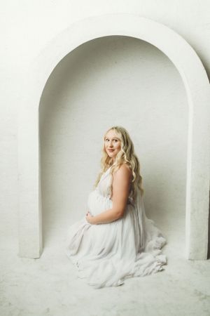 Maternity photoshoot featuring a woman in white dress posing in an alcove.