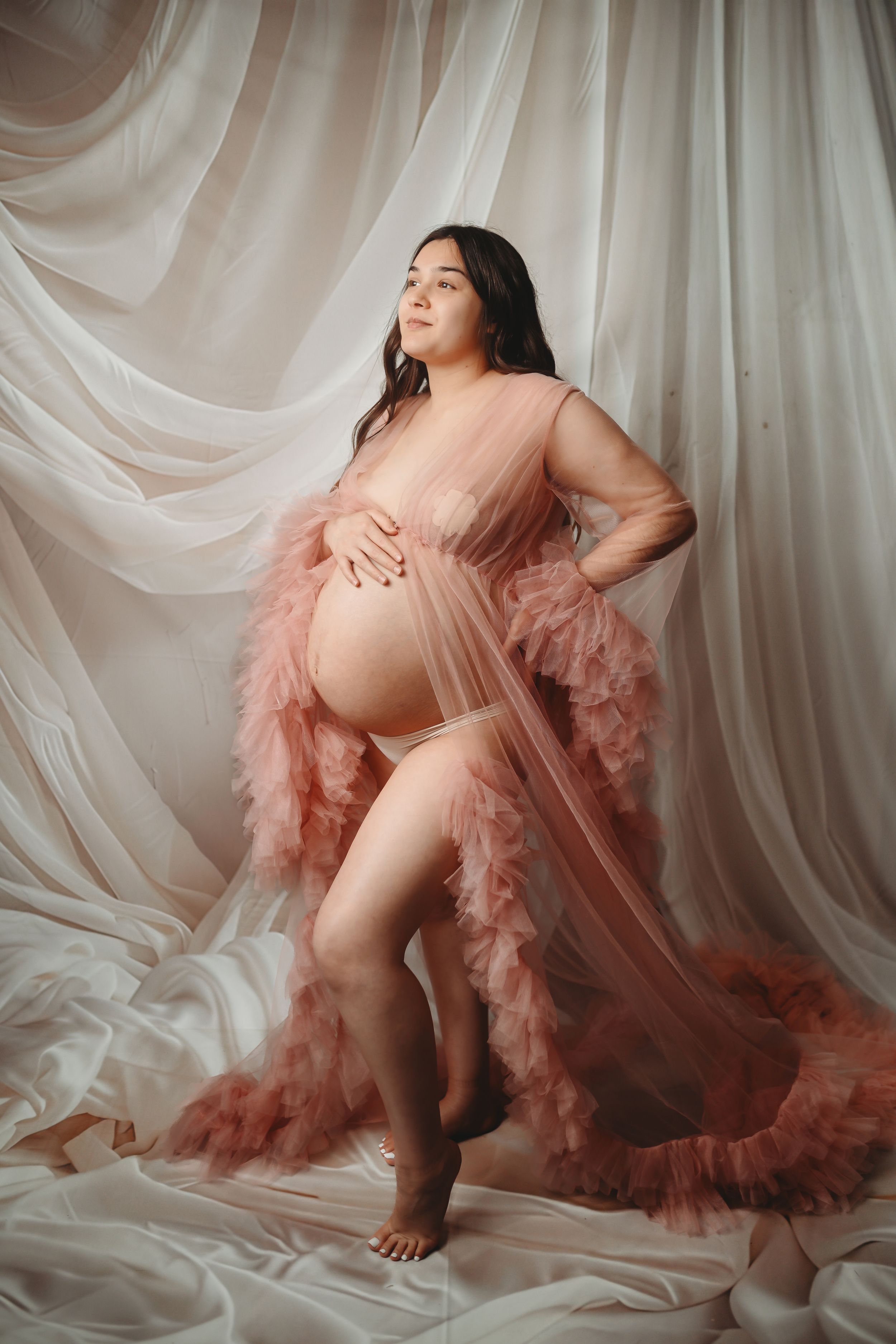 Maternity photo with flowing pink tulle dress against white fabric backdrop.