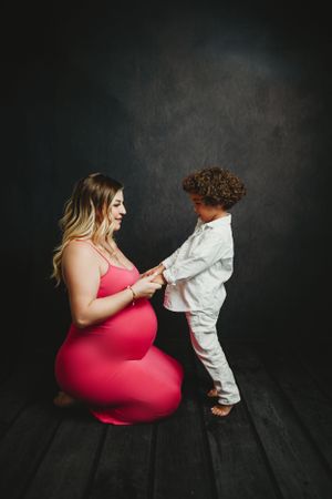 Maternity session with woman in pink dress and child in white shirt against dark backdrop.