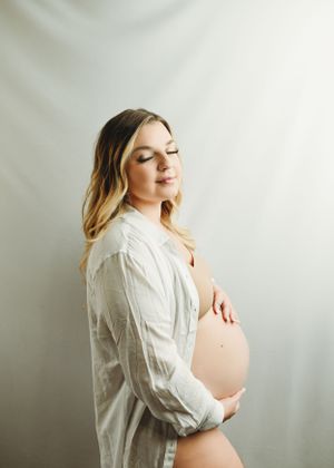 Artistic maternity portraits in white showing expectant mother in flowing shirt against light backdrop.
