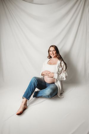 Relaxed maternity poses on white backdrop wearing casual denim and white shirt.