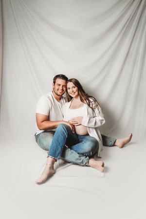 A couple in casual attire sits together on a white backdrop in a cozy studio portrait session.