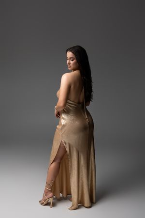 Woman in backless gold satin gown with slit, posing elegantly in strappy heels against gray background.