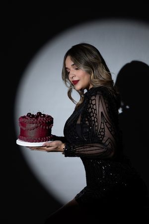 Elegant figure in black lace dress holds red velvet cake in dramatic spotlight.