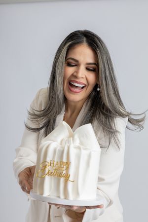 Person in white outfit joyfully presents birthday cake with gold lettering.