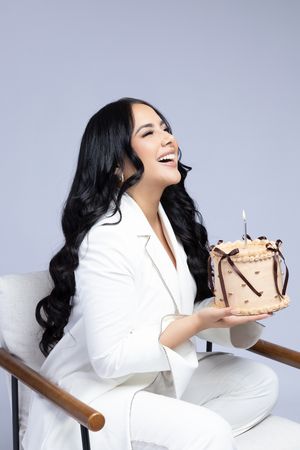 Person in white suit smiles while holding birthday cake with candle.