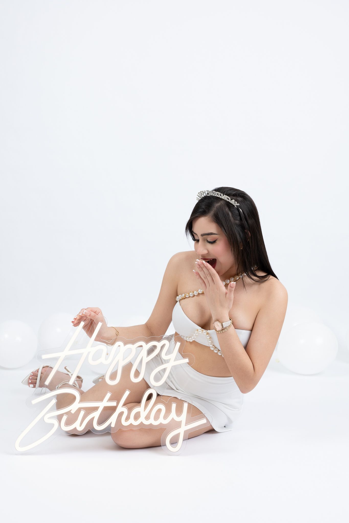 Woman in white holds an illuminated Happy Birthday neon sign against a bright white background.