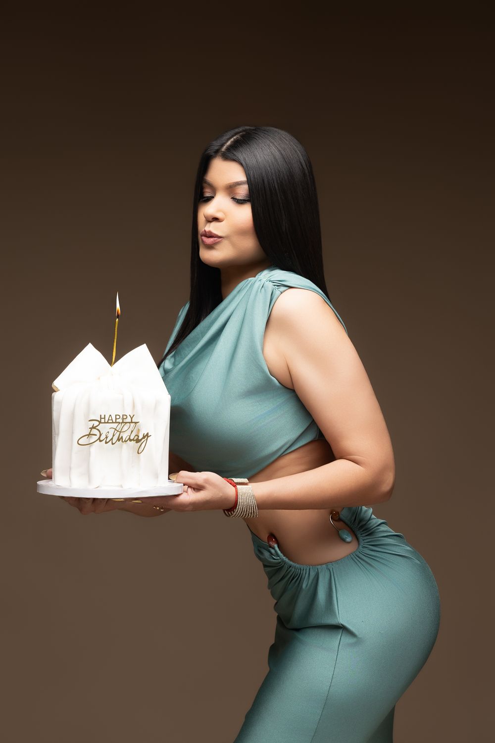 Woman in teal outfit holding a white Happy Birthday cake with a lit candle on a brown background.