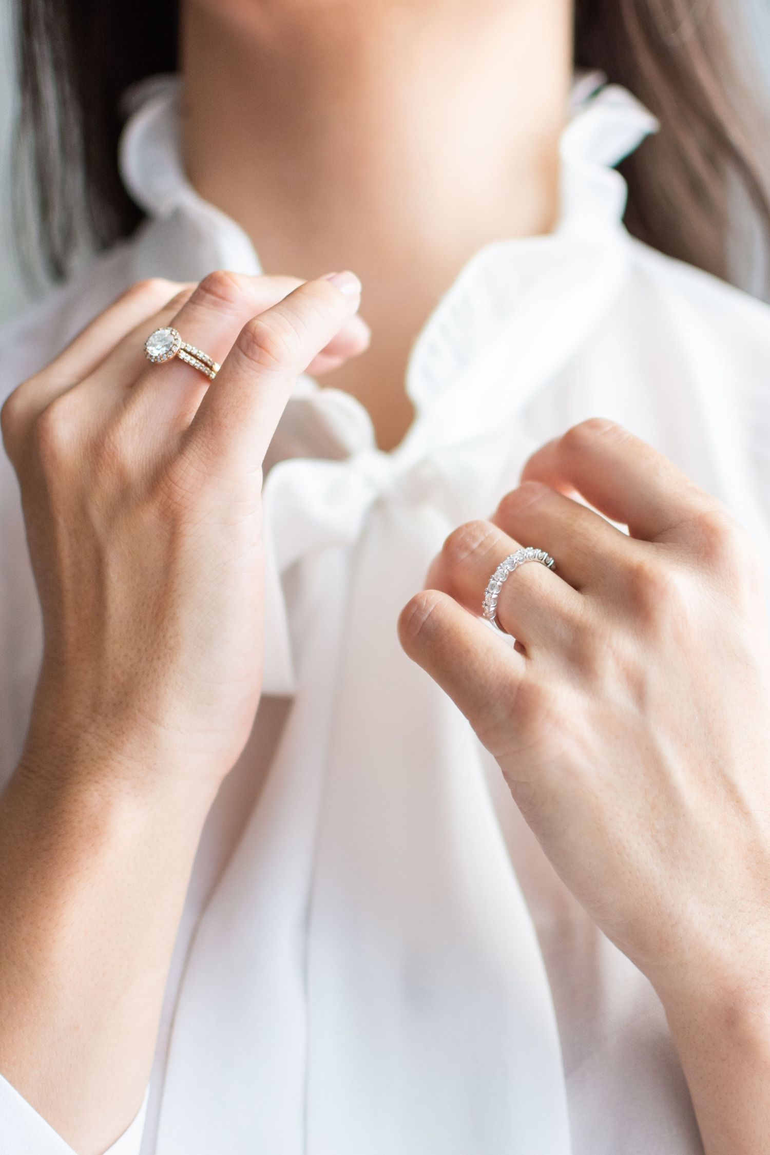 Close up view of hands wearing engagement rings against white clothing background.