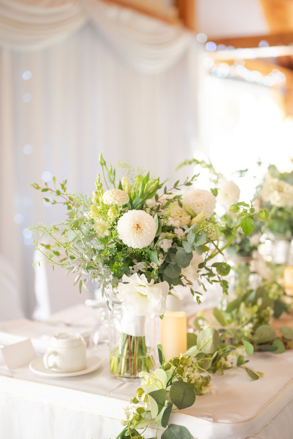 Elegant white floral centerpiece with garden roses and greenery on wedding reception table with soft lighting and candles.