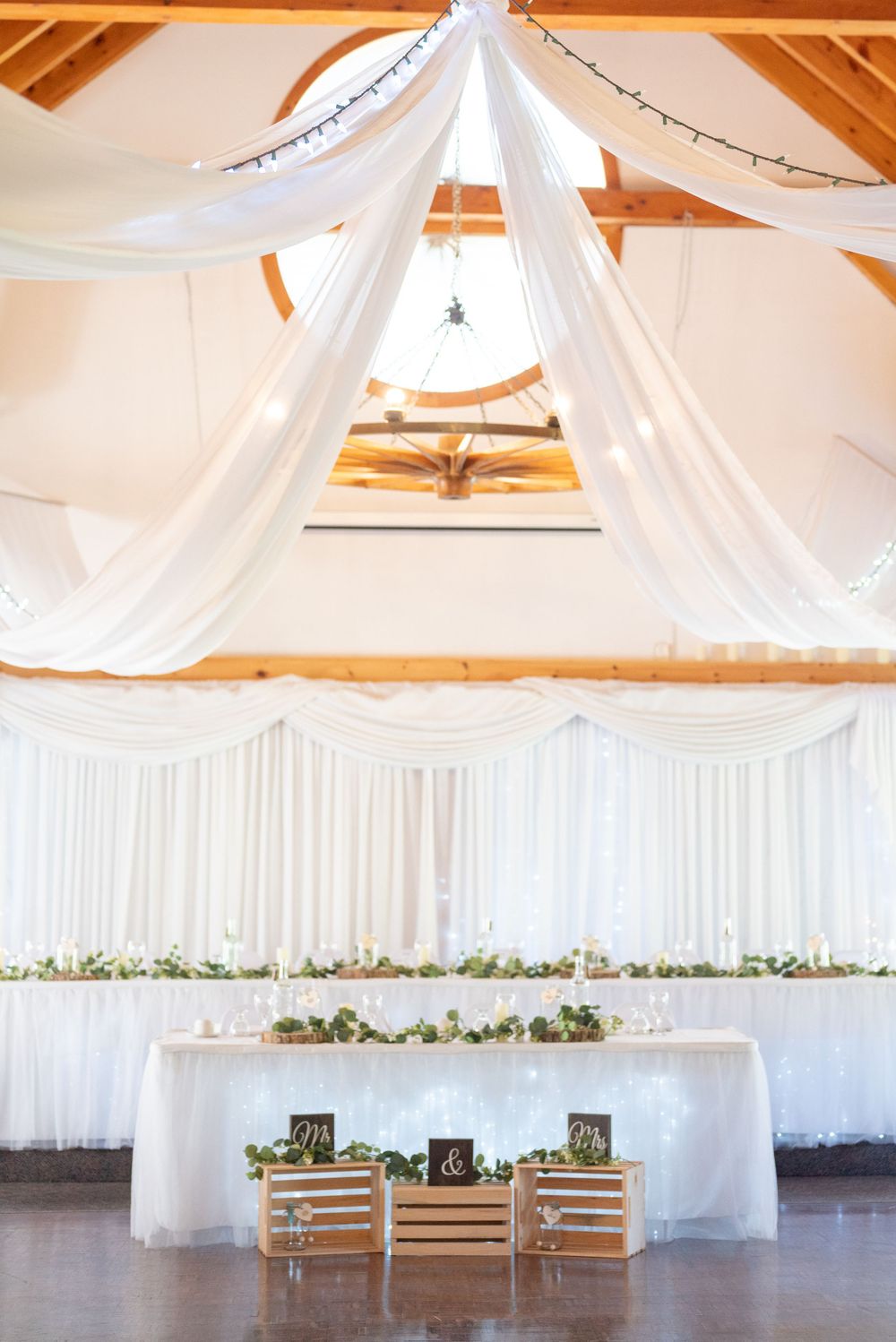 White draping and floral decor create an elegant wedding reception setup with rustic wooden chairs at a head table.
