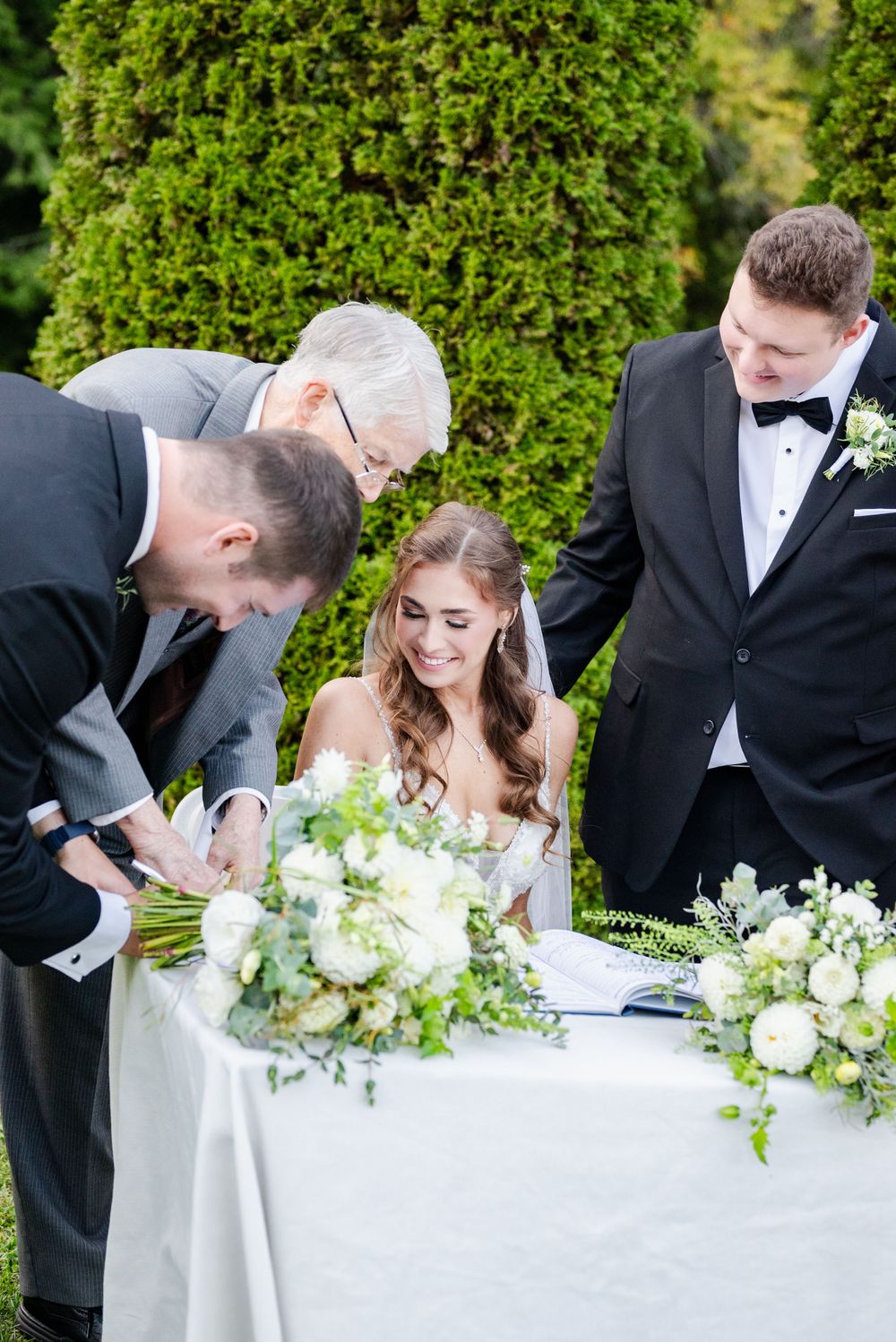 People gather at an elegant outdoor wedding ceremony around a white-clothed table with lush floral arrangements.
