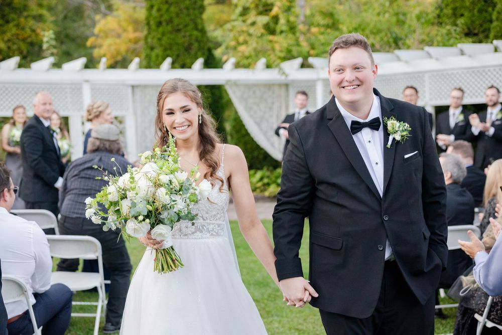 Newlyweds walk hand in hand down the aisle at outdoor garden ceremony with white floral bouquet and classic attire.