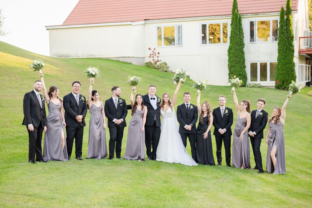 Wedding party members in formal attire stand in a line on a green hillside with a white building in the background.
