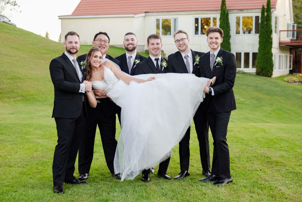 A bride in a white wedding gown is lifted up by groomsmen in black suits on a grassy hillside.