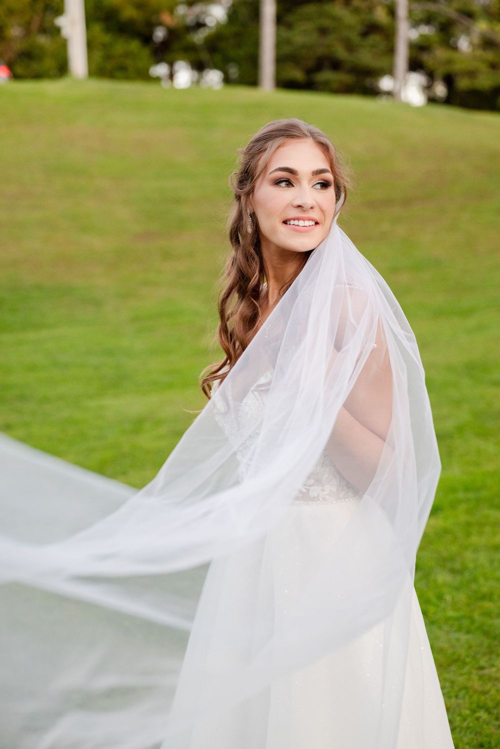 A bride in a flowing white veil and wedding dress poses outdoors on a green lawn during a sunny day.