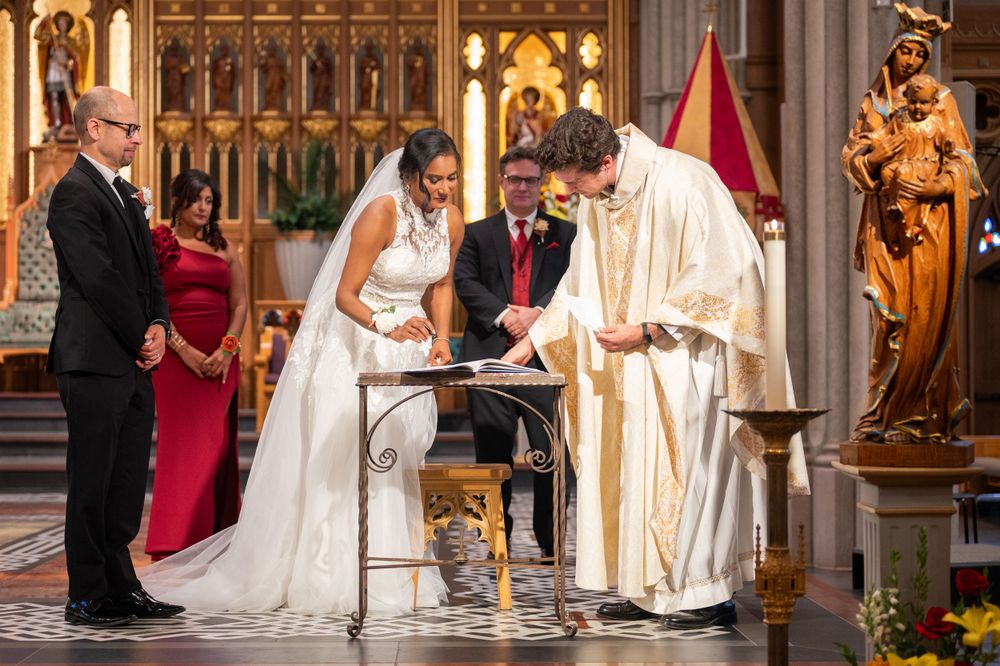 A couple signs their marriage register at the altar with a priest and witnesses in an ornate church setting.
