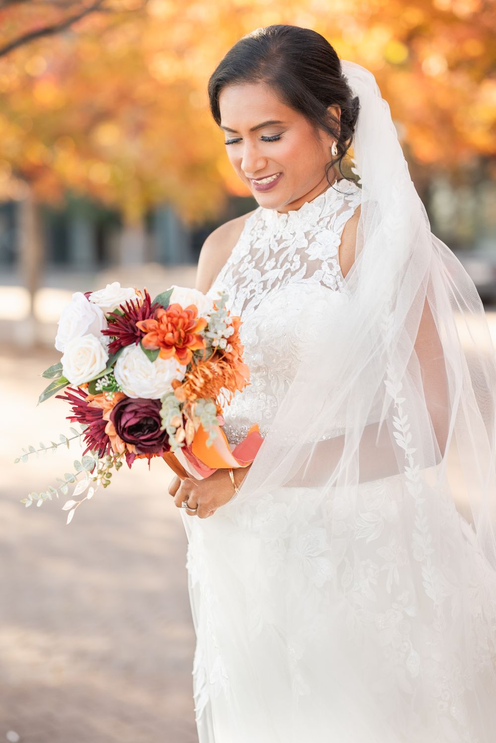 Bride in lace gown holds vibrant fall bouquet with burgundy and orange flowers against autumn foliage background.