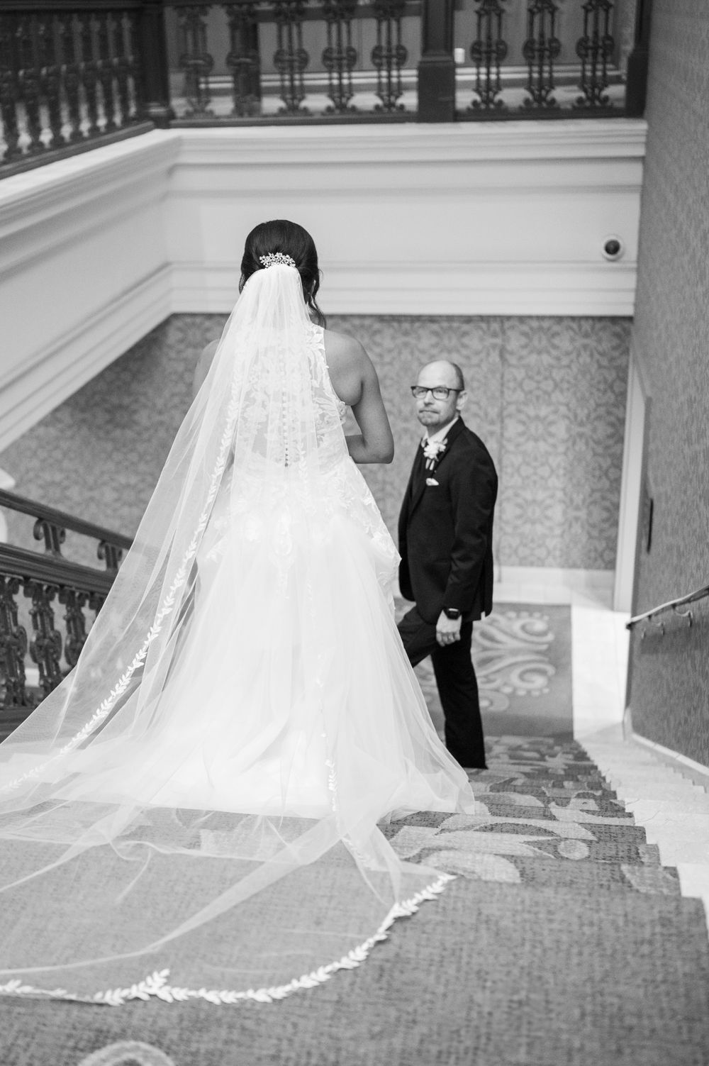 Black and white wedding photo showing bride in flowing veil descending elegant staircase with floral carpet.