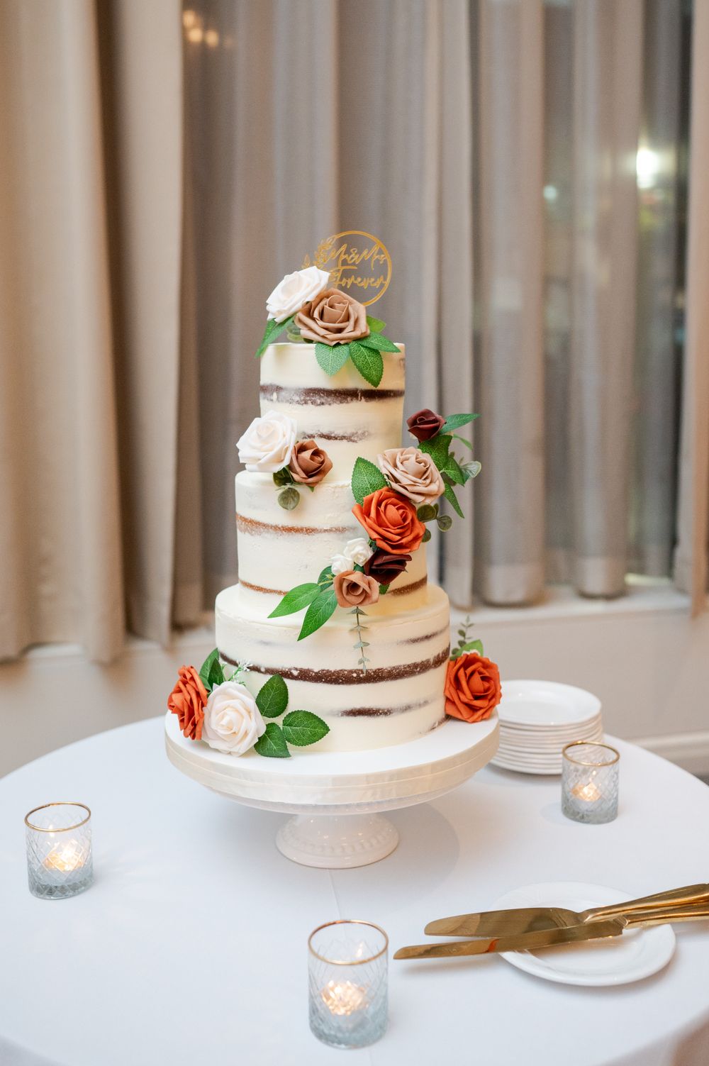 Elegant three-tier semi-naked wedding cake decorated with orange roses and greenery on white cake stand with votive candles.