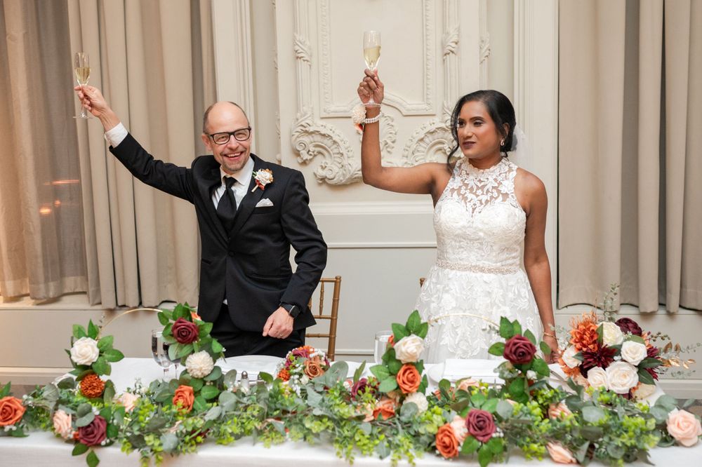 A bride and groom raise their hands joyfully at their reception table decorated with lush floral arrangements.