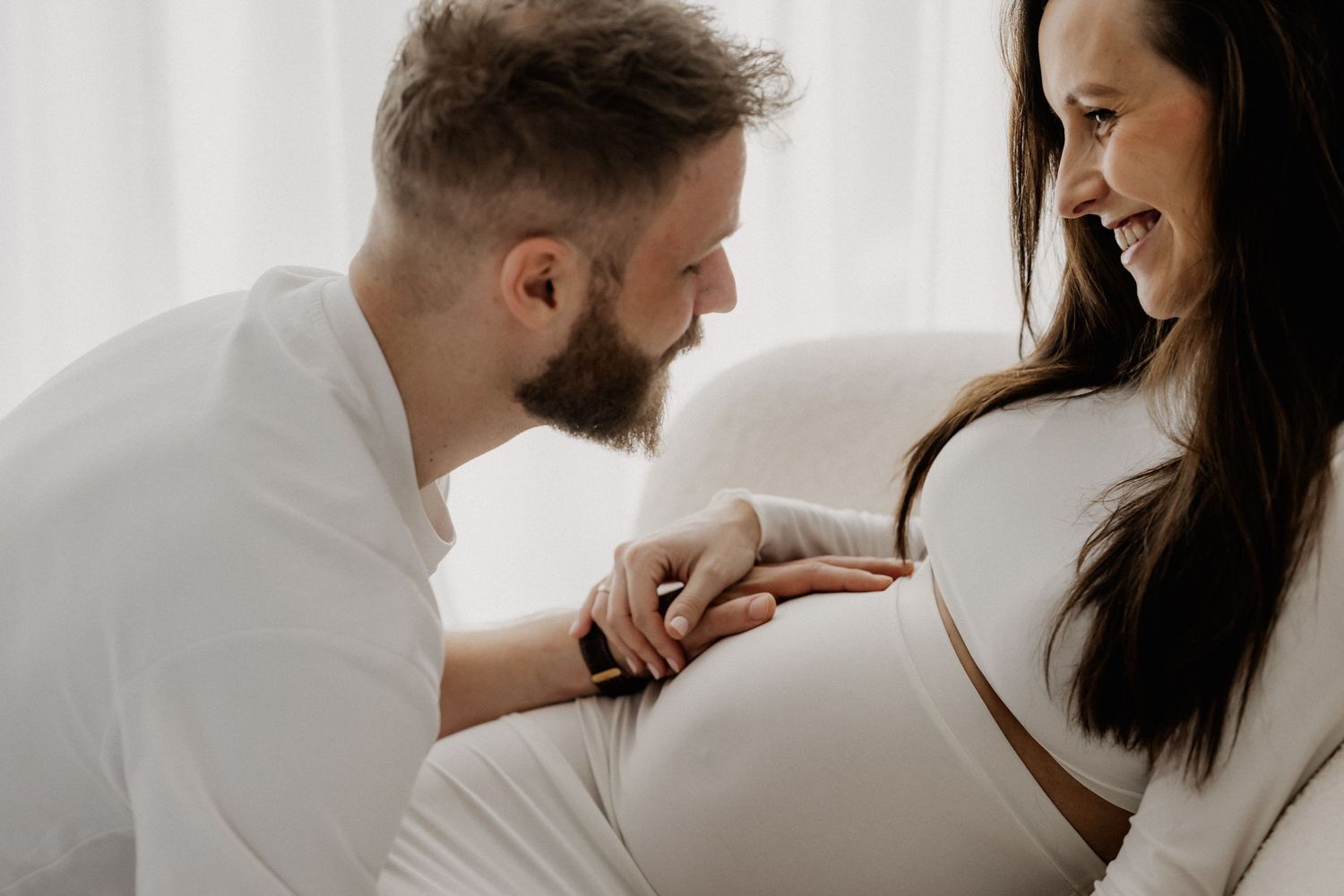 Werdende Eltern teilen einen zärtlichen Moment, der Mann beugt sich vor, um den Bauch der schwangeren Frau zu küssen, in weicher weißer Umge