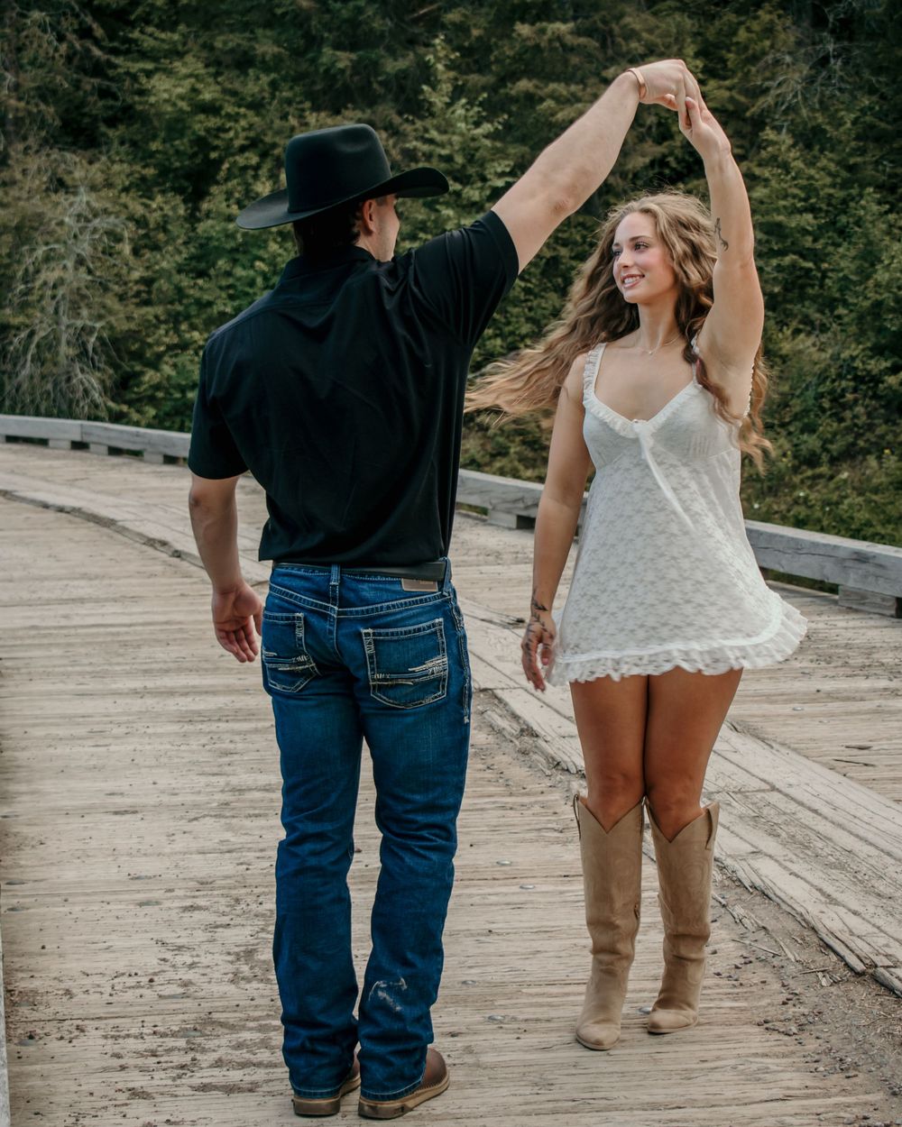 A couple dancing together on a wooden bridge, with one wearing a black cowboy hat and the other in a white dress.