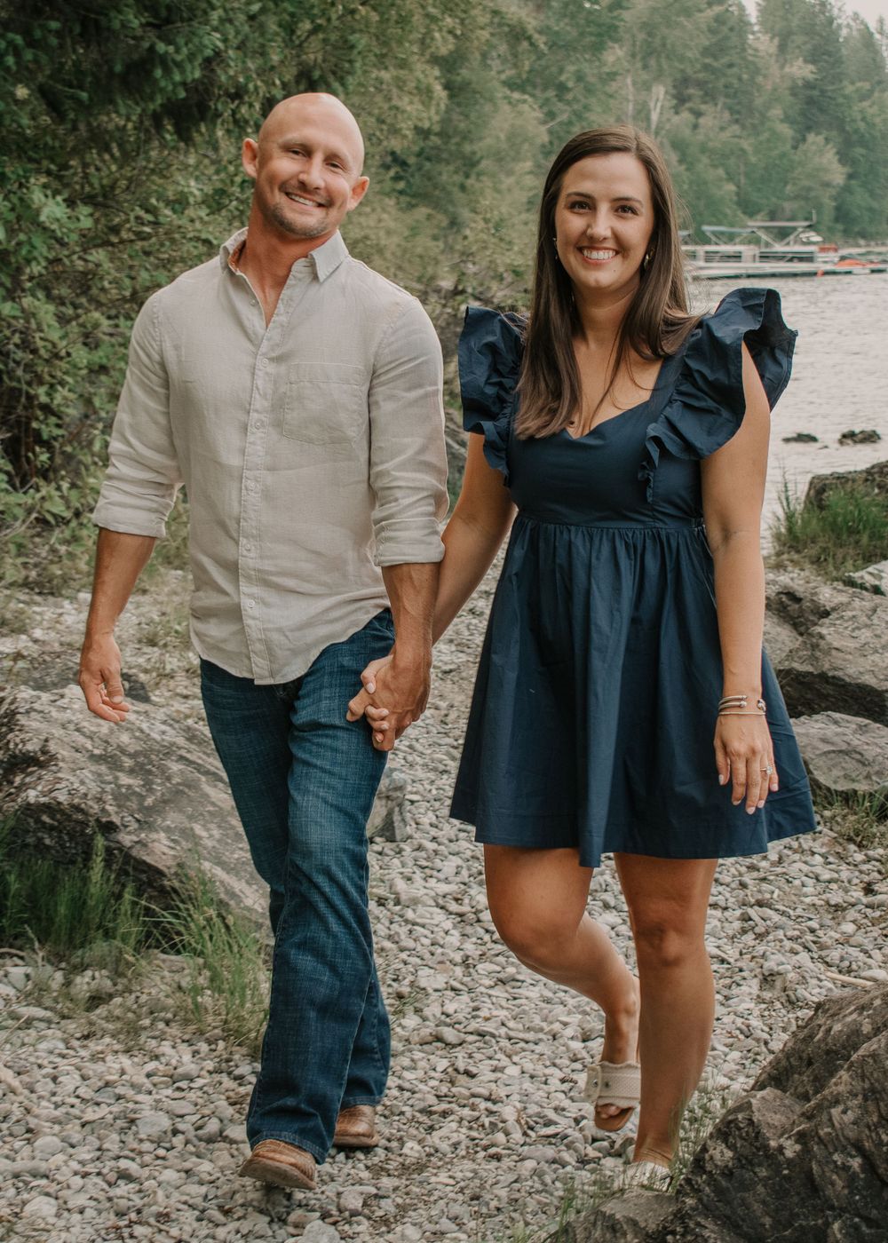 Couple in casual attire walking along a rocky shoreline by a lake.