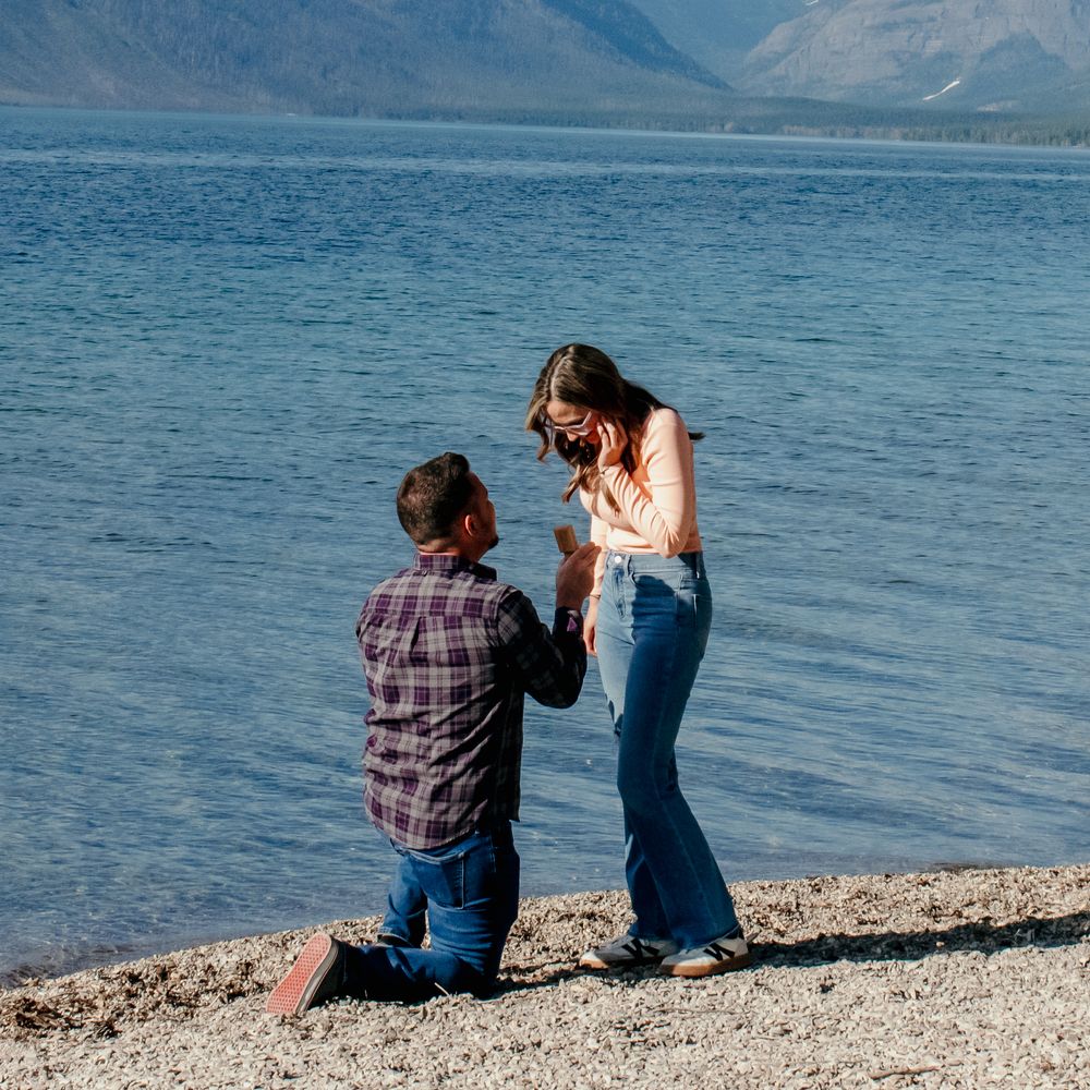 Two people share a special moment sitting on a rocky beach shore with mountains and lake in the background.