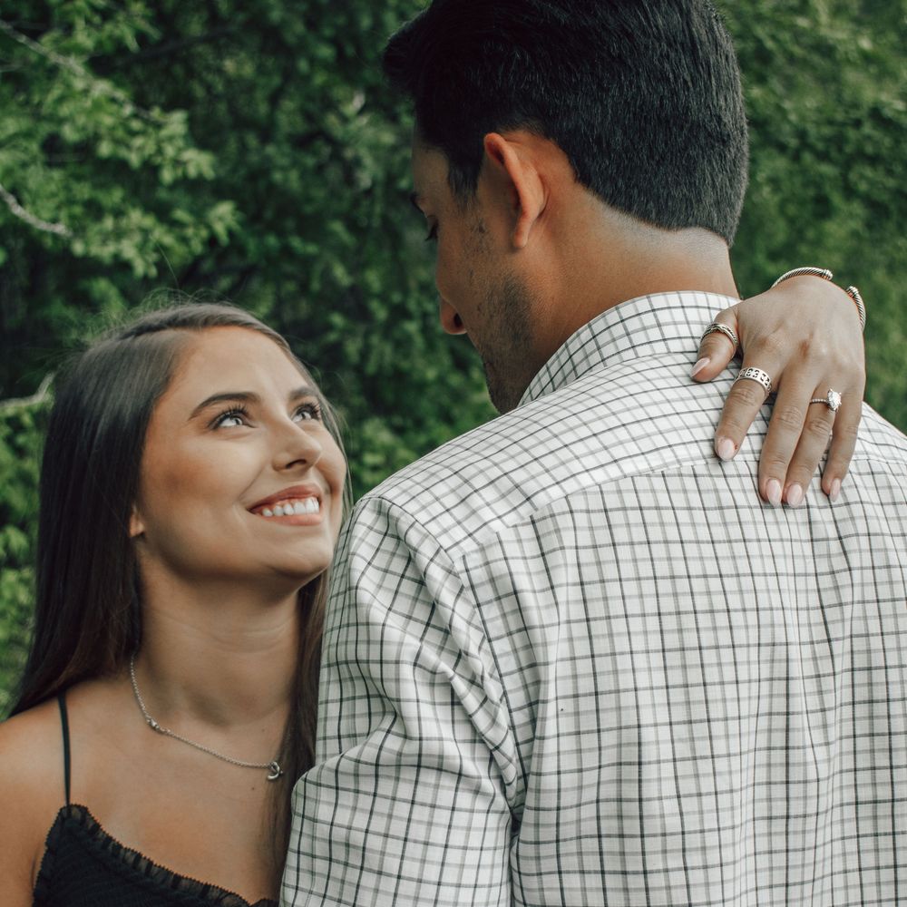 A couple embraces outdoors, with the woman smiling lovingly while resting her hand on her partner's shoulder.