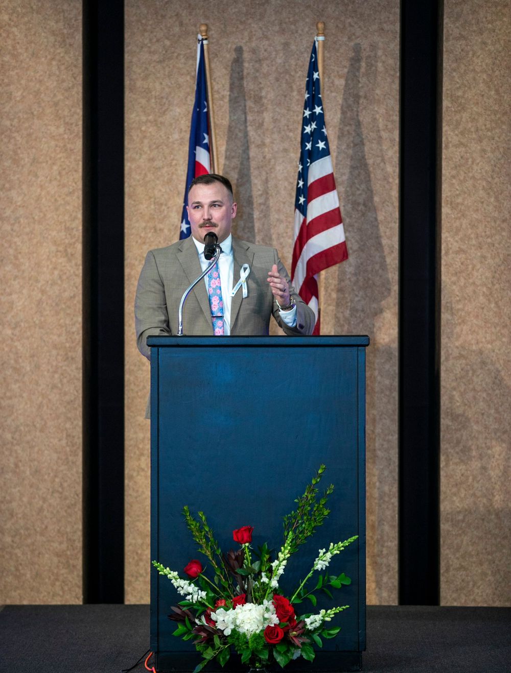 Man in suit speaking at podium with American flag backdrop at formal event.