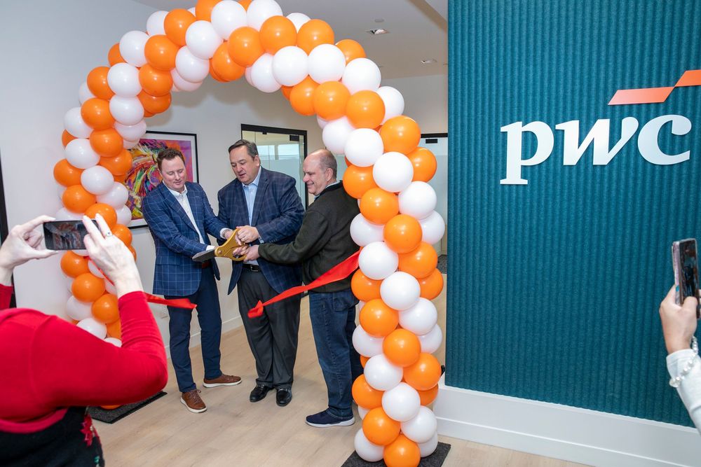 Three men cut a red ribbon at a PwC office grand opening ceremony under an orange and white balloon arch.