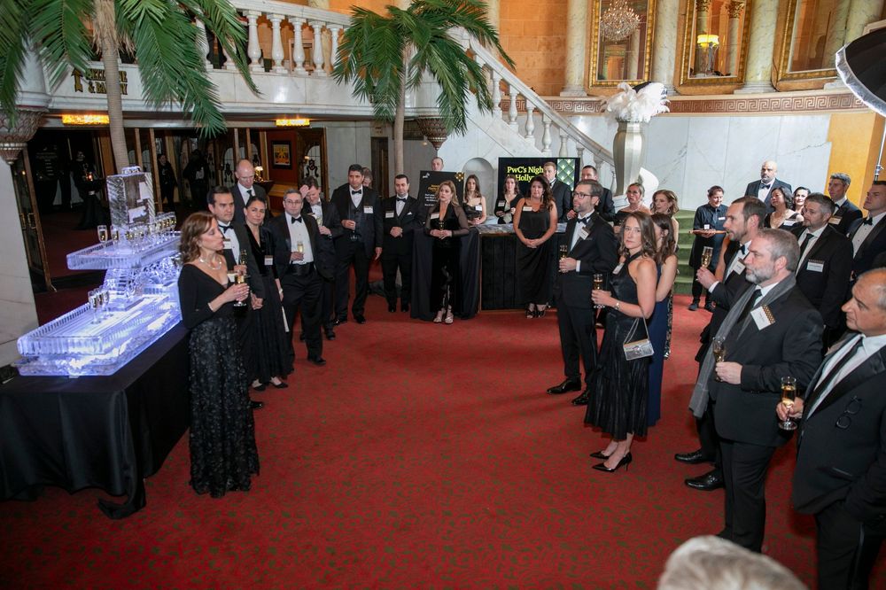 Elegantly dressed guests mingle at a formal gala event in an ornate venue with red carpet and ice sculpture.