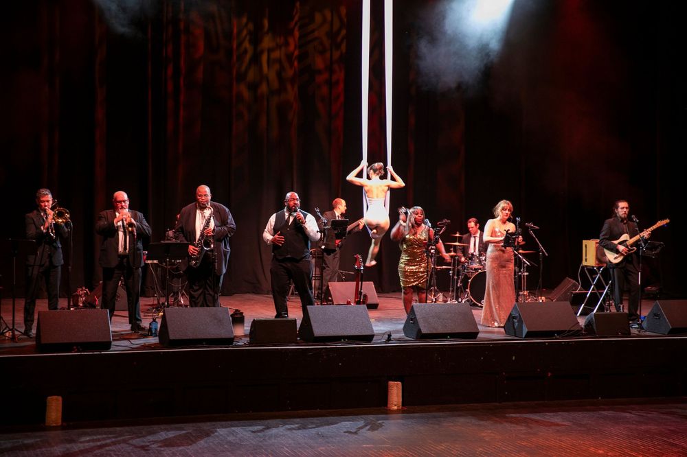 A large band performs on a lit stage with an aerial acrobat suspended above them during a live show.
