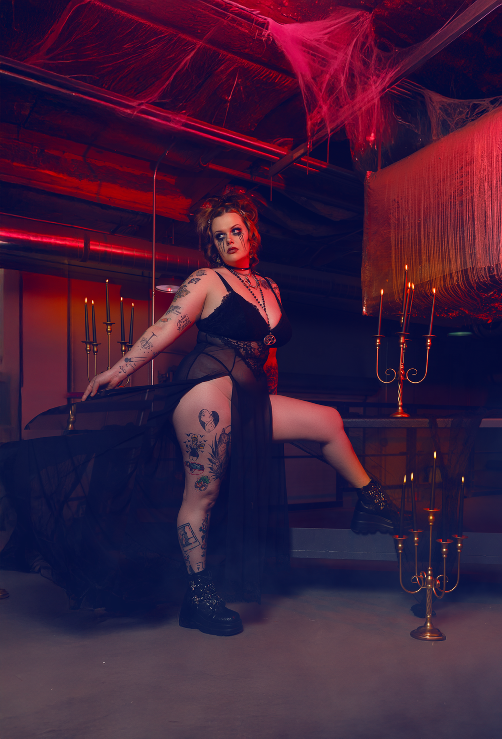 Gothic figure poses dramatically by candelabra in red-lit industrial space.