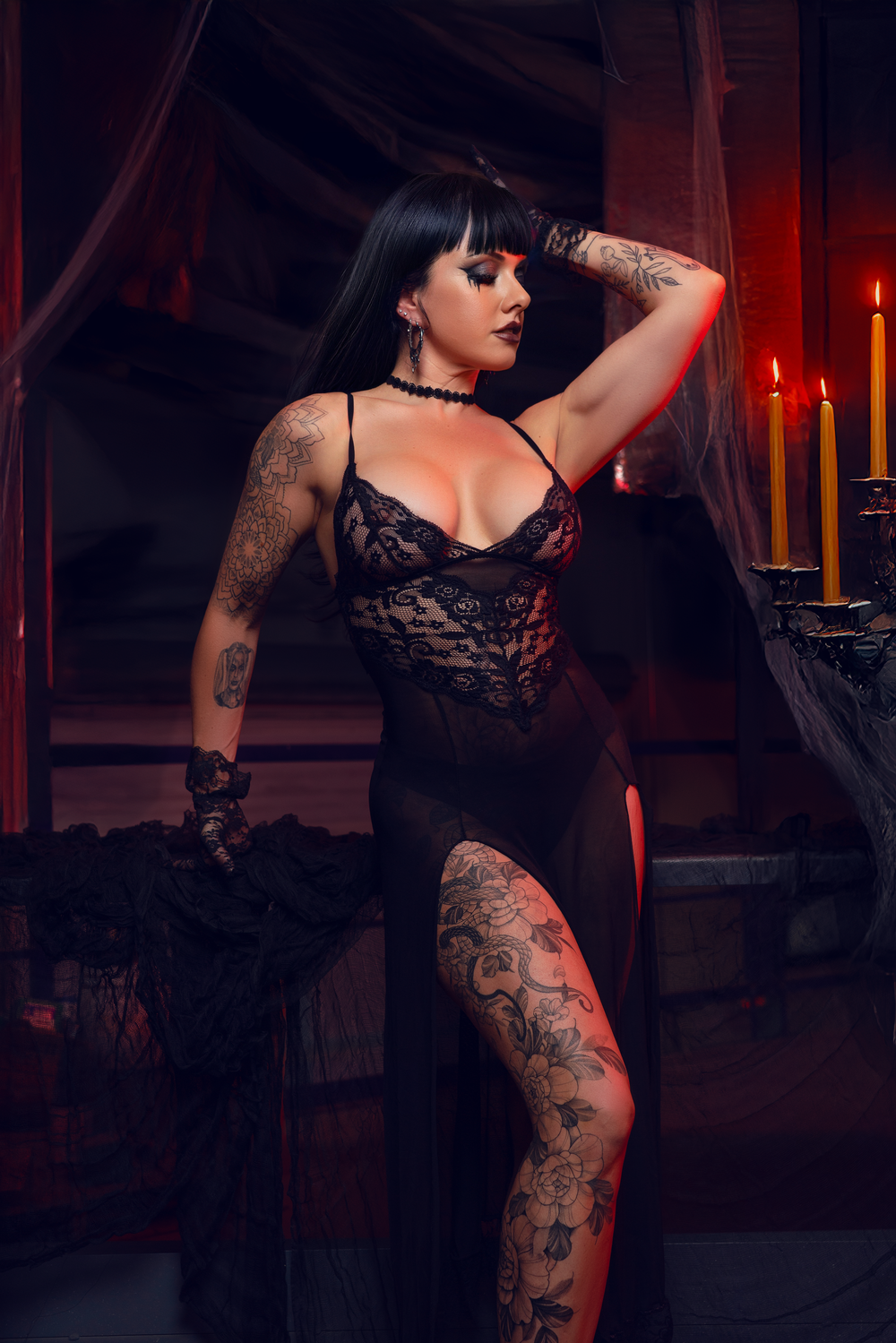 Gothic model in black lace dress poses with tattoos and candles in dark atmospheric setting.