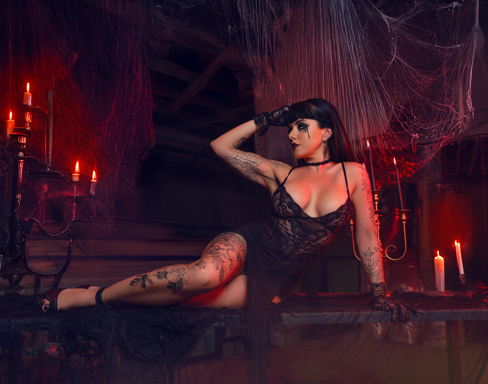 Dark figure reclines on floor in gothic setting with red candles and cobwebs.