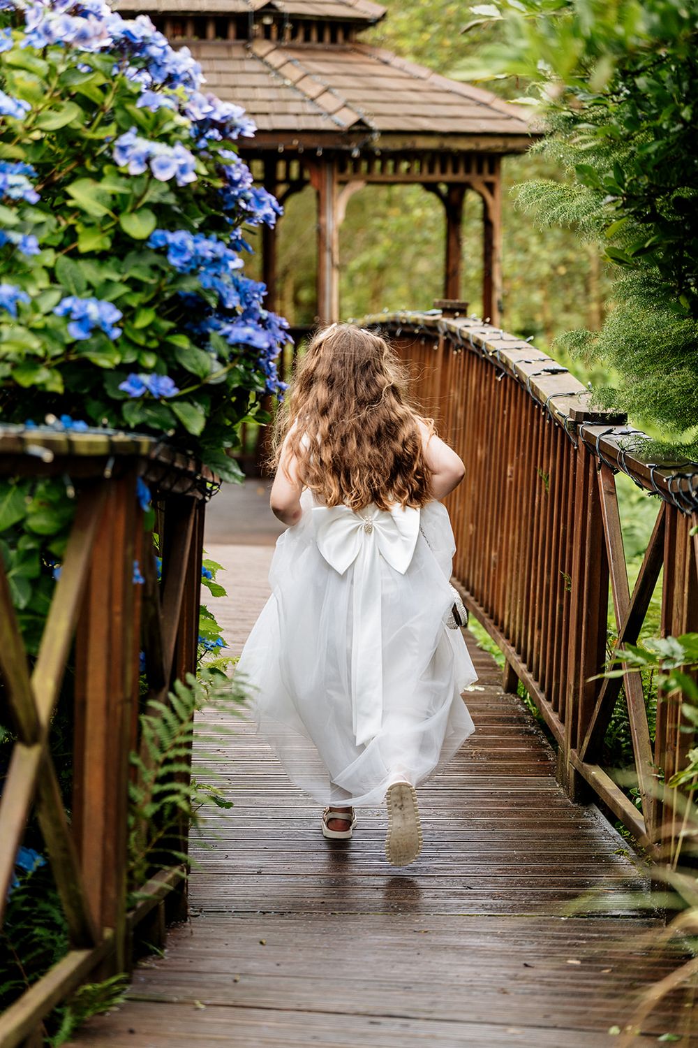 A small figure in a white dress with bow detail walks along a wooden bridge path near blooming blue hydrangeas and a gazebo.