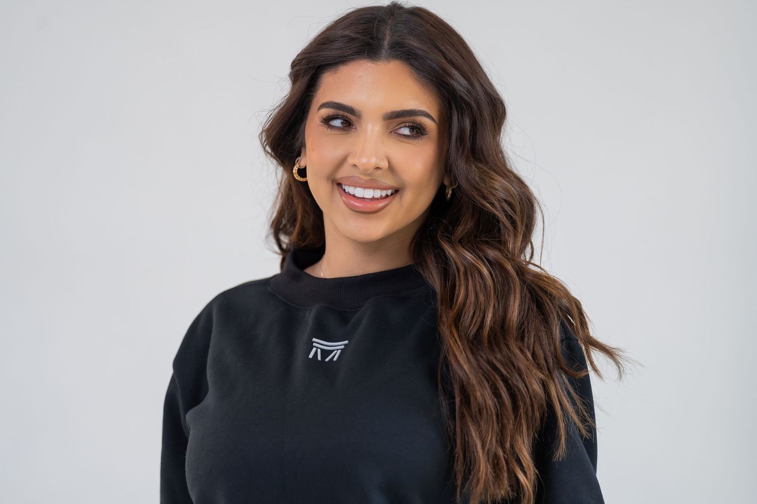 Smiling young woman with long wavy brown hair wearing a black sweatshirt, posing against a light gray background.
