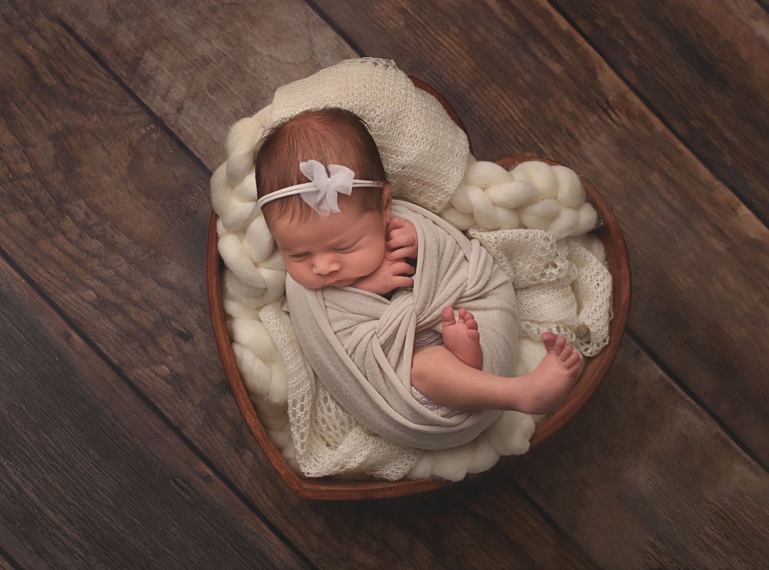 Sleeping newborn wrapped in white fabric and headband rests peacefully in heart-shaped basket on wooden floor.