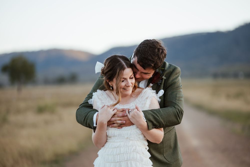 Groom in green shirt embracing bride in white ruffled dress on rural dirt road with mountain backdrop.