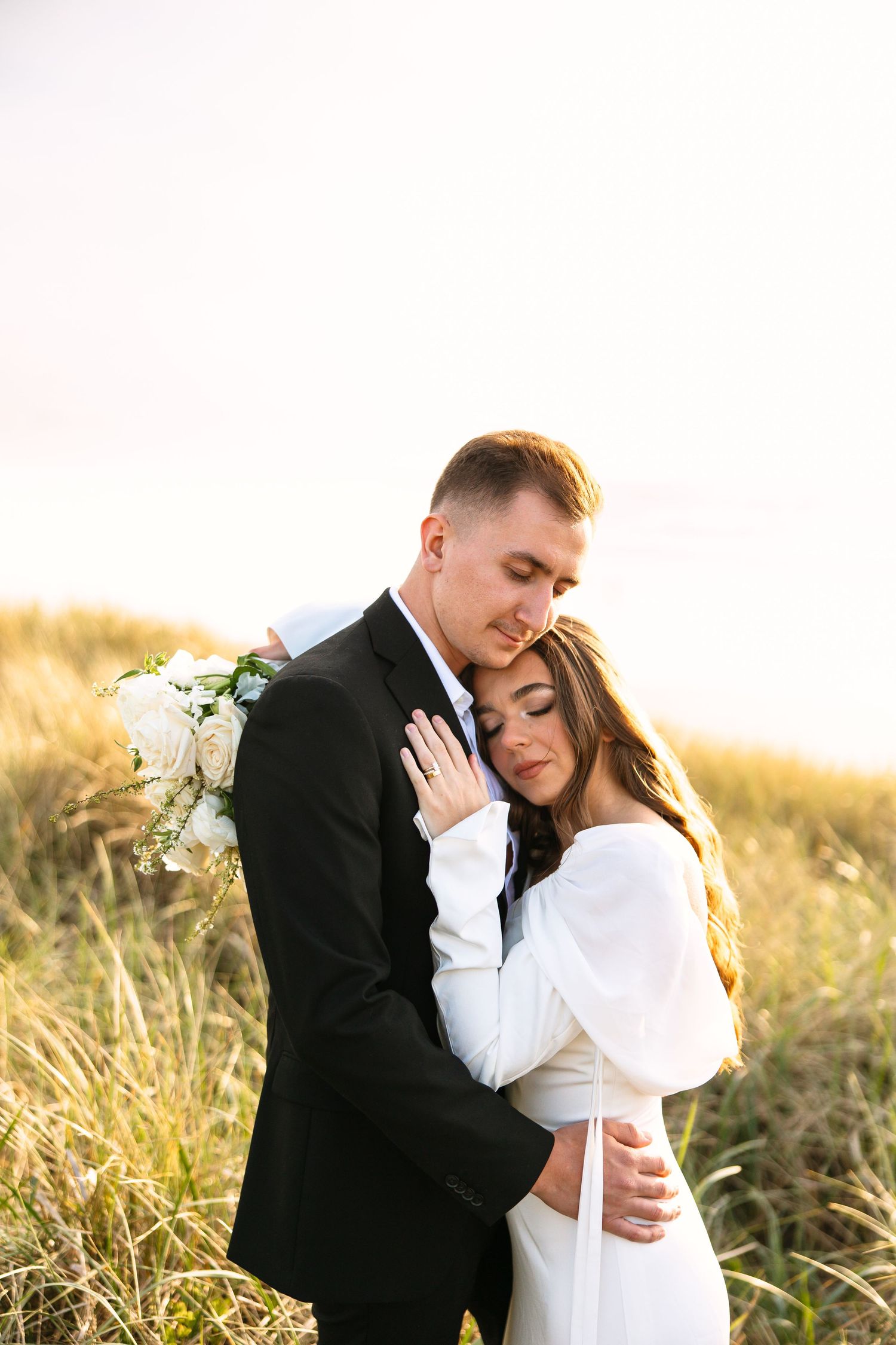 A tender wedding portrait in golden light showing an embrace among tall grass.
