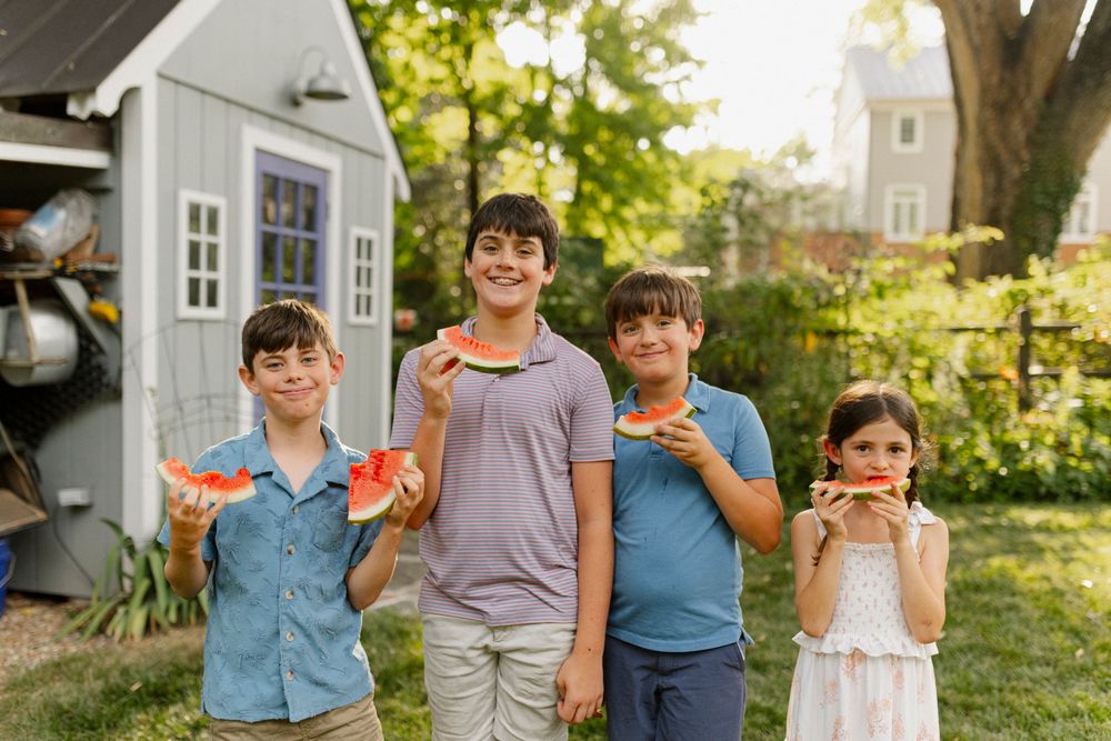 Four children smiling and holding watermelon slices in a sunny backyard with a gray shed behind them.