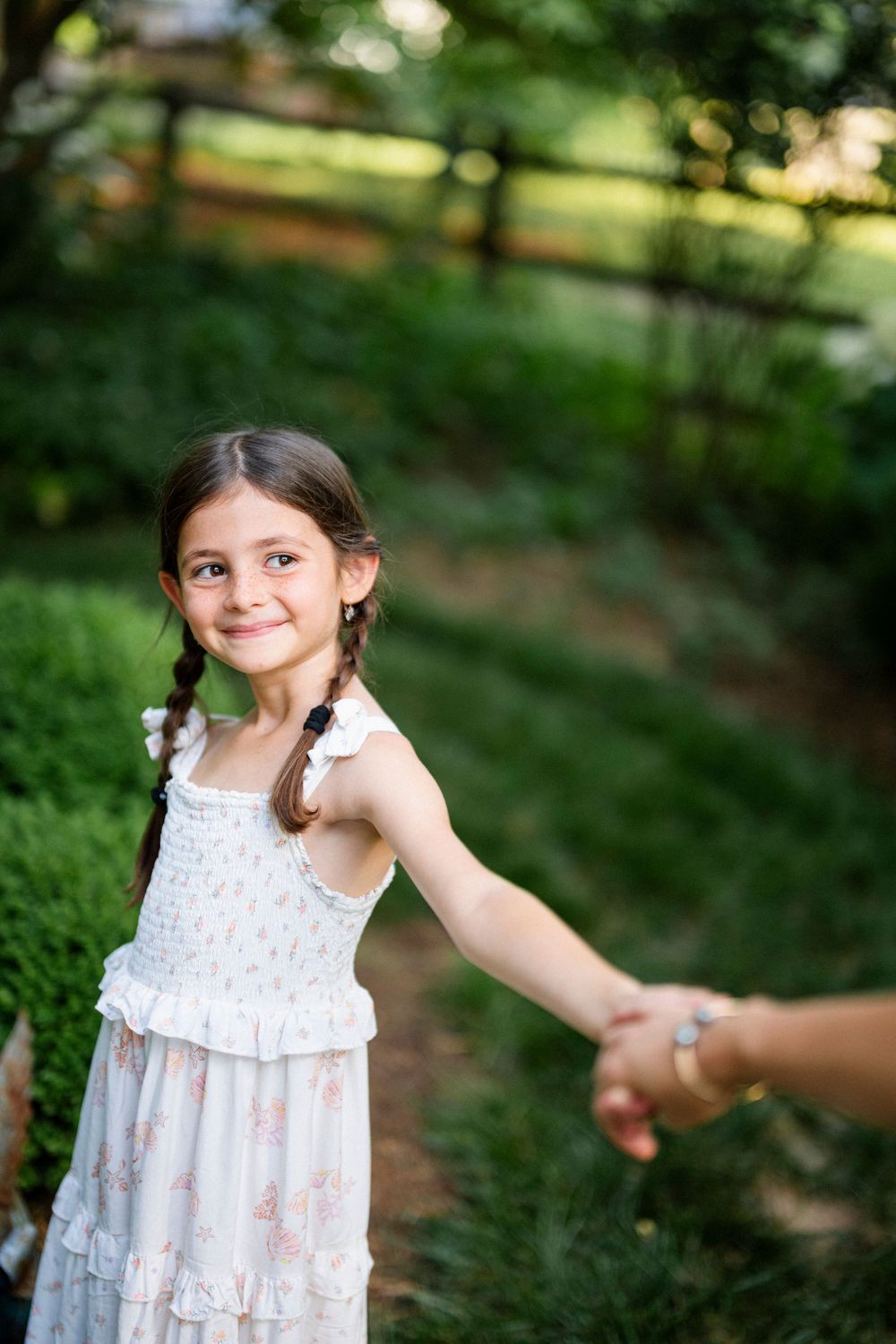 A young girl in a white floral dress with braids smiles while holding an adult's hand in a lush green park.